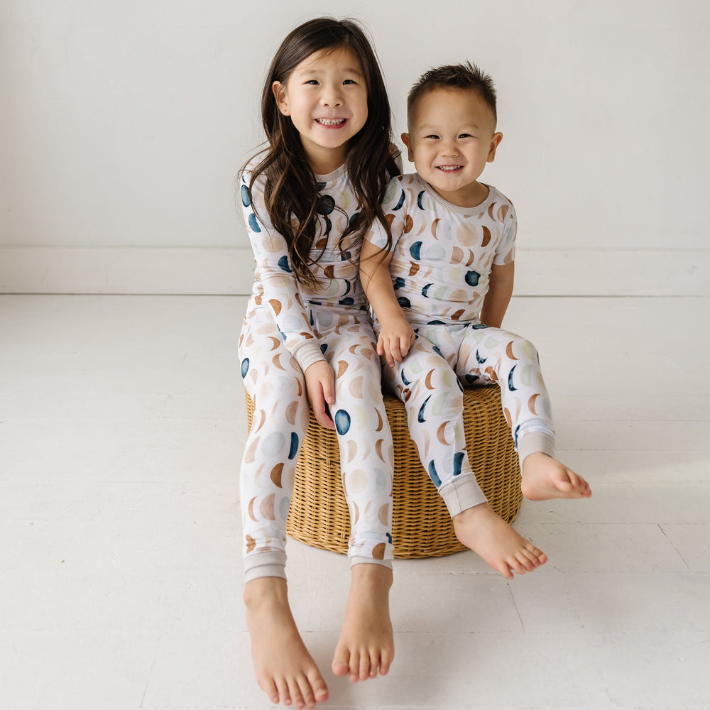 Image of two toddlers in pajama sets in Luna Neutral print. This print features phases of the moon in the sweetest shades of creams, tans, and navy watercolor in an all over repeat pattern.