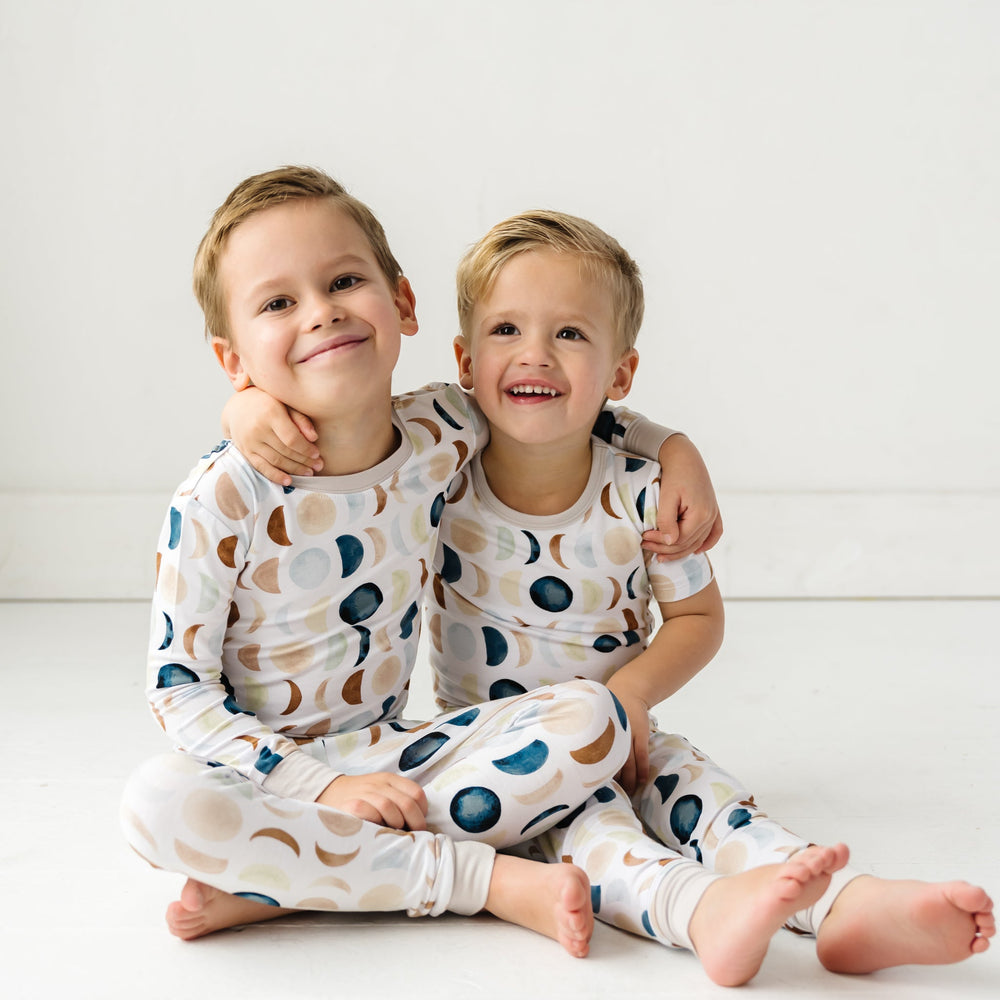 Image of two toddlers in pajama sets in Luna Neutral print. This print features phases of the moon in the sweetest shades of creams, tans, and navy watercolor in an all over repeat pattern.