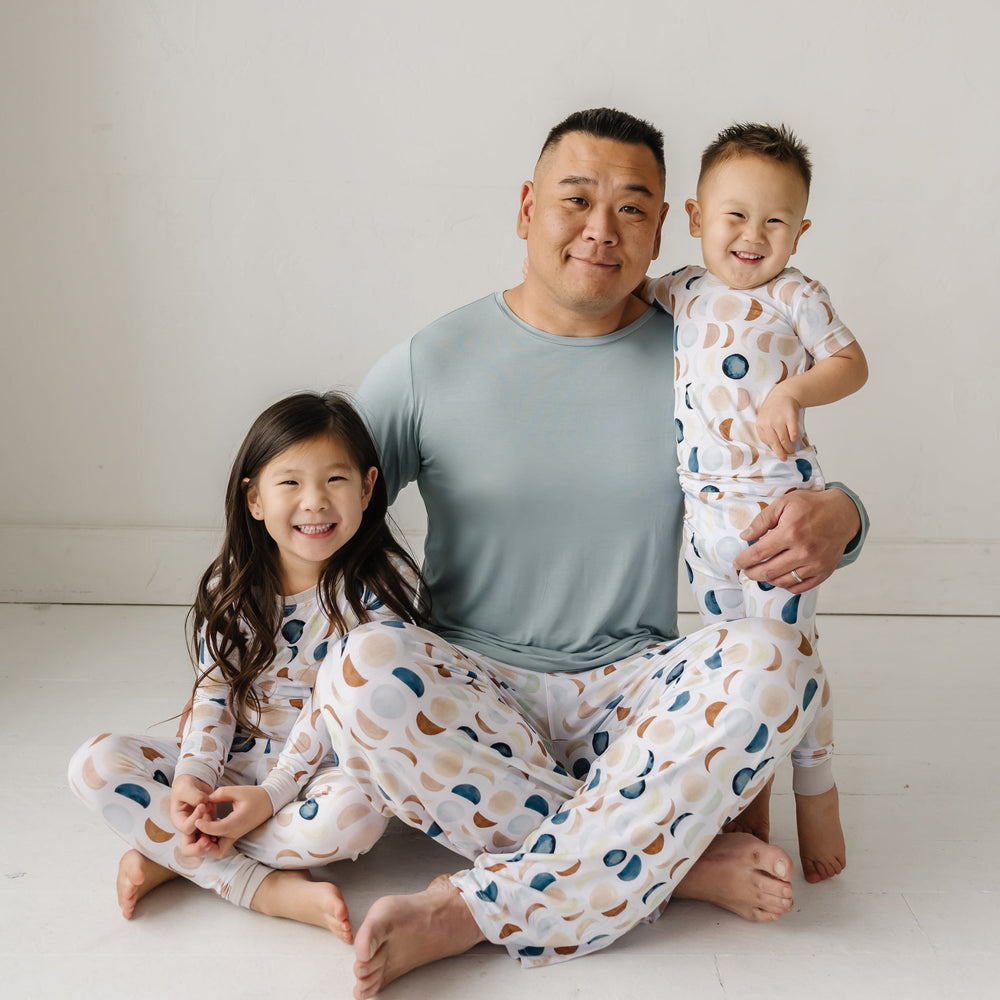 Image of a father in Luna Neutral printed pajama pants paired with solid Stormy colored top and two children in matching printed pajamas. This print features phases of the moon in the sweetest shades of creams, tans, and navy watercolor in an all over repeat pattern.