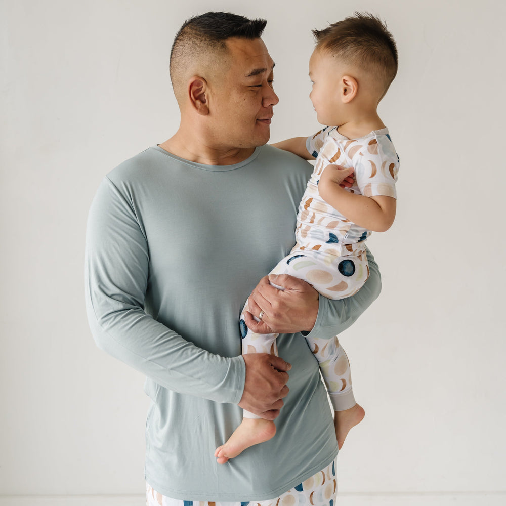 Image of a father in Luna Neutral printed pajama pants paired with solid Stormy colored top and one child in matching printed pajamas. This print features phases of the moon in the sweetest shades of creams, tans, and navy watercolor in an all over repeat pattern.
