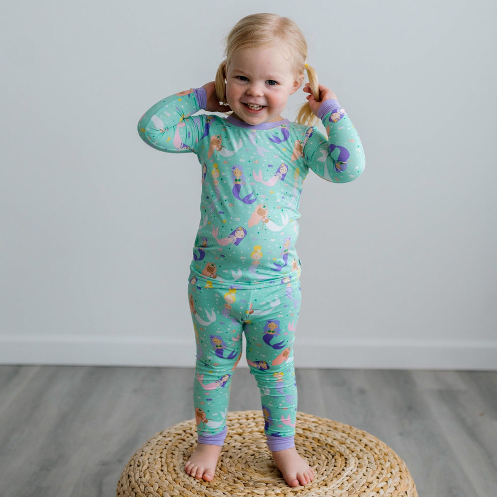 Image of toddler girl posing on a rattan pouf. She is shown wearing a mermaid printed two-piece pajama set. This print includes multi-colored mermaids and fish that are featured on an aqua background with a purple trim. 