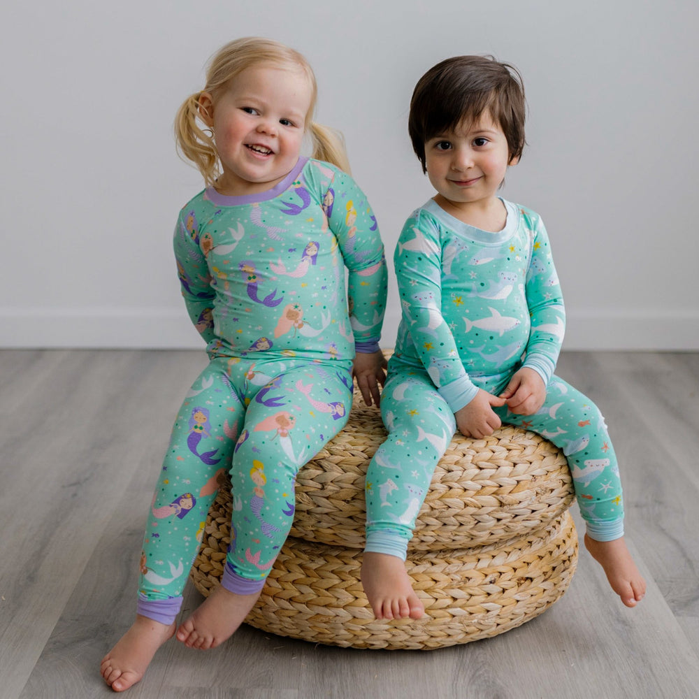 Image of toddler girl and boy sitting on a rattan pouf. The girl is shown wearing a mermaid printed two-piece pajama set while the boy is shown wearing a shark printed two-piece pajama set. 