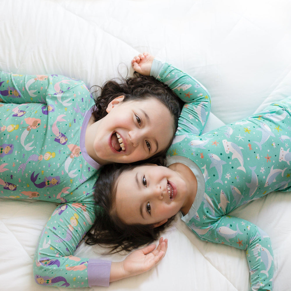 Click to see full screen - Image of two little girls laying on a bed. One is wearing a mermaid printed two-piece pajama set while the other is in a shark printed two-piece pajama set. 