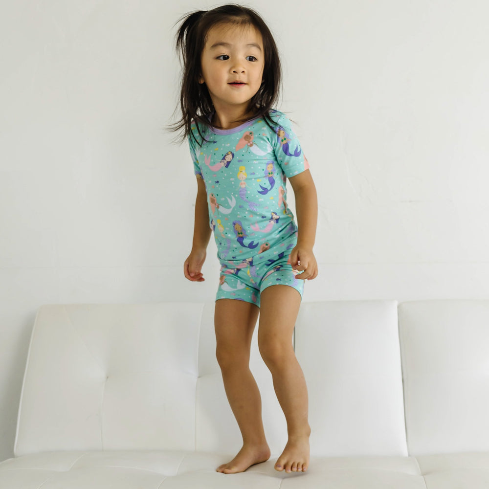 Click to see full screen - Image of little girl wearing a mermaid printed short sleeve and shorts pajama set. Image of infant girl wearing a mermaid printed short sleeve and shorts zip up romper. This print includes multi-colored mermaids and fish that are featured on an aqua backg