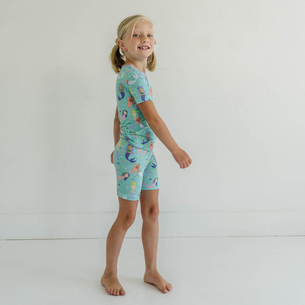 Click to see full screen - Image of little girl wearing a mermaid printed short sleeve and shorts pajama set. Image of infant girl wearing a mermaid printed short sleeve and shorts zip up romper. This print includes multi-colored mermaids and fish that are featured on an aqua backg