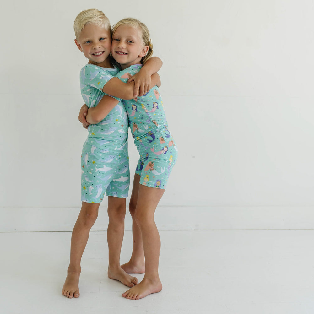 Image of little boy and girl wearing coordinating short sleeve and shorts pajama sets. The boy is shown wearing a shark printed pj set, while the girl is shown in a mermaid printed set. This print includes multi-colored mermaids and fish that are featured