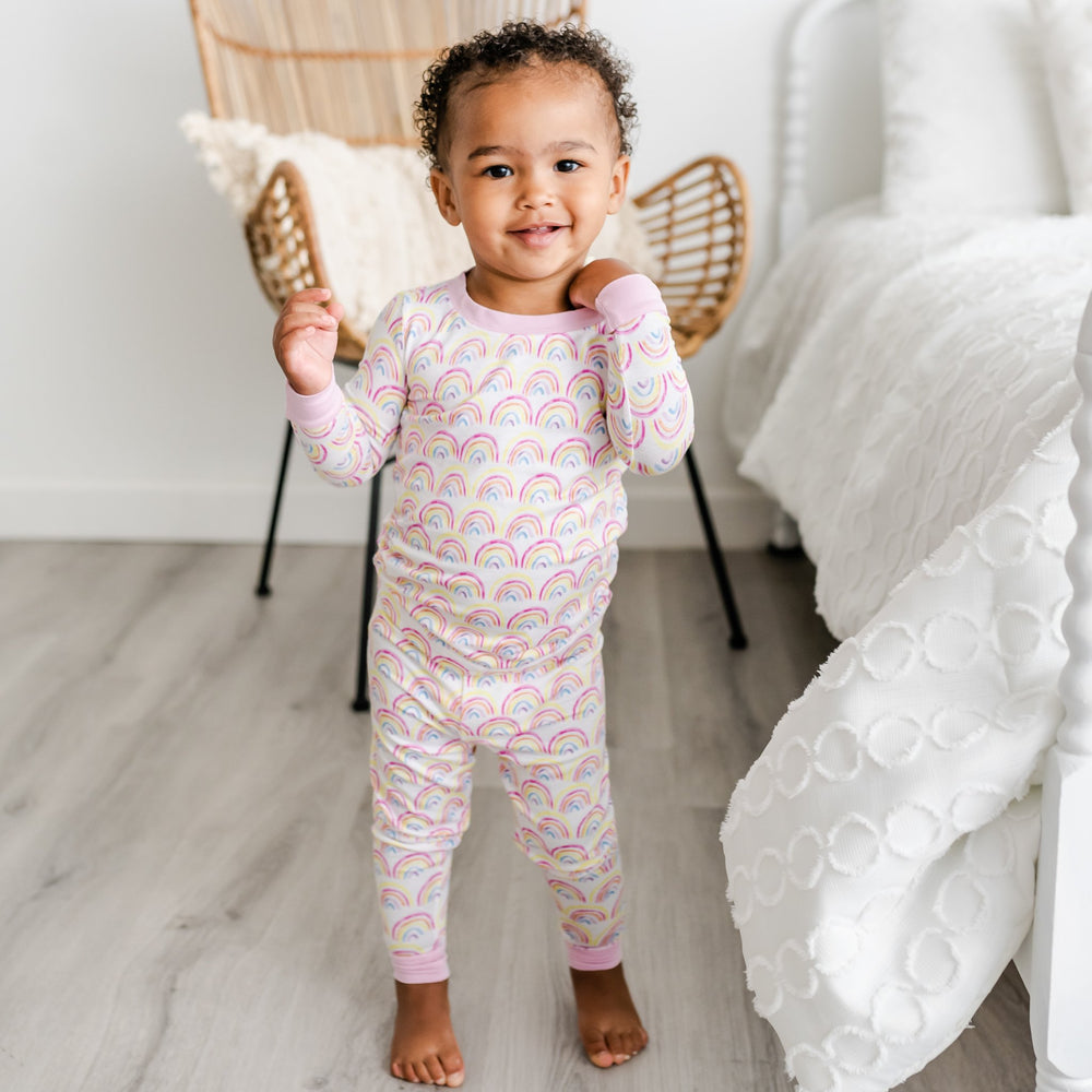 Image of toddler girl wearing two-piece pajama set in rainbow print. This print features multicolored rainbows that sit upon a white background with light pink accent trim.