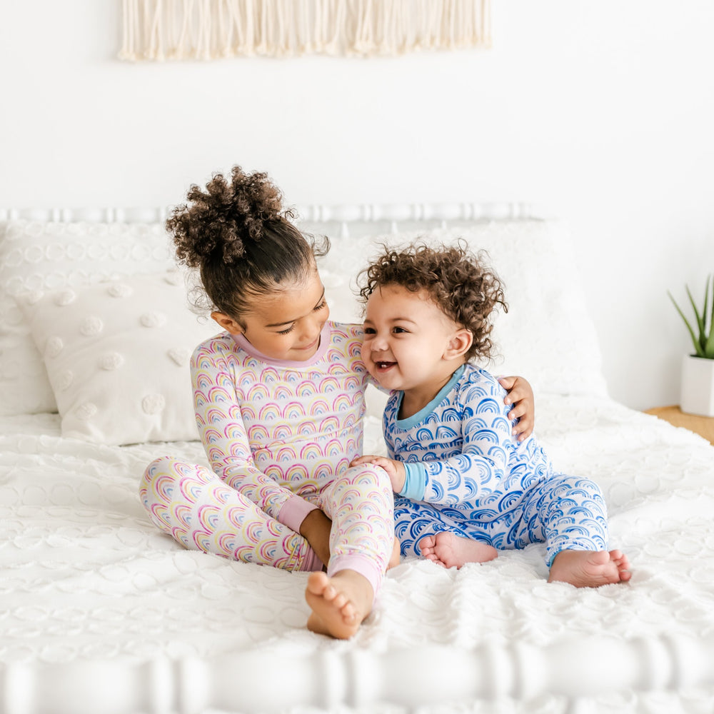 Image of toddler girl and boy wearing matching rainbow printed pajama sets. Toddler girl is shown wearing pink rainbow pajama set and toddler boy is shown wearing blue rainbow pajama set. 