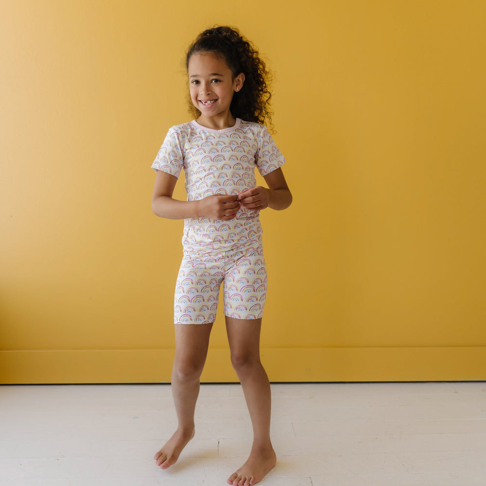 Image of little girl wearing a rainbow printed short sleeve and shorts pajama set. This print features multicolored rainbows that sit upon a white background with light pink accent trim.