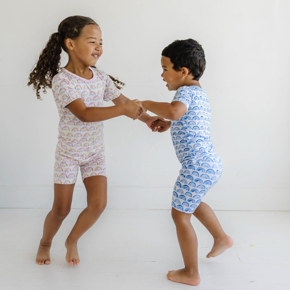 Image of little girl and boy holding hands, they are shown wearing coordinating rainbow printed short sleeve and shorts pajama sets. The girl is shown wearing the pink rainbow print, and the boy is shown wearing blue. 
