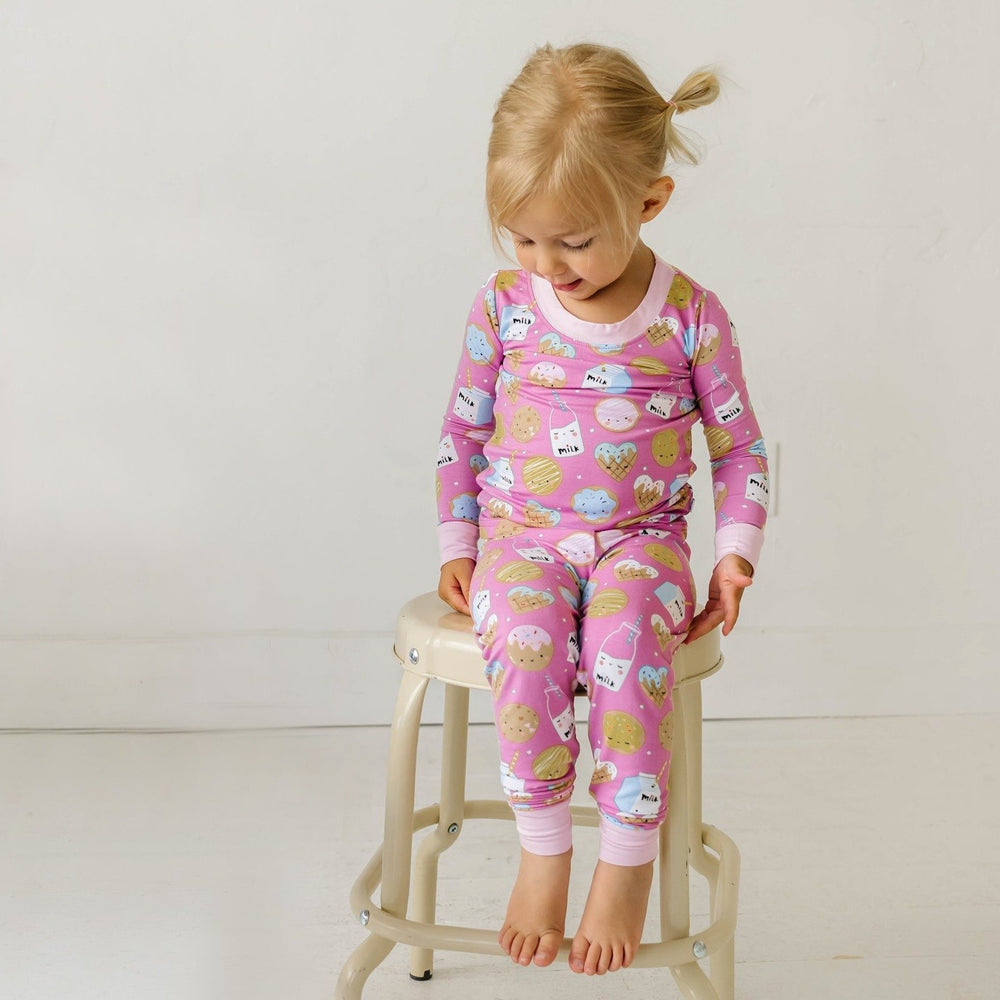 Image of little girl sitting on a stool. She is shown wearing a two-piece pajama set in cookies and milk print. This print features milk cartons, colorful sprinkled cookies, and chocolate chip cookies that sit upon a pink background with light pink trim. 