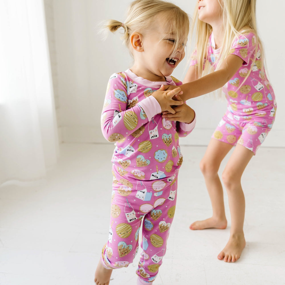 Image of two little girls chasing each other. They are shown wearing matching pajamas in cookies and milk print. One little girl is shown wearing a two-piece pajama set, while the other is shown wearing a short sleeve and shorts pajama set. This print fea