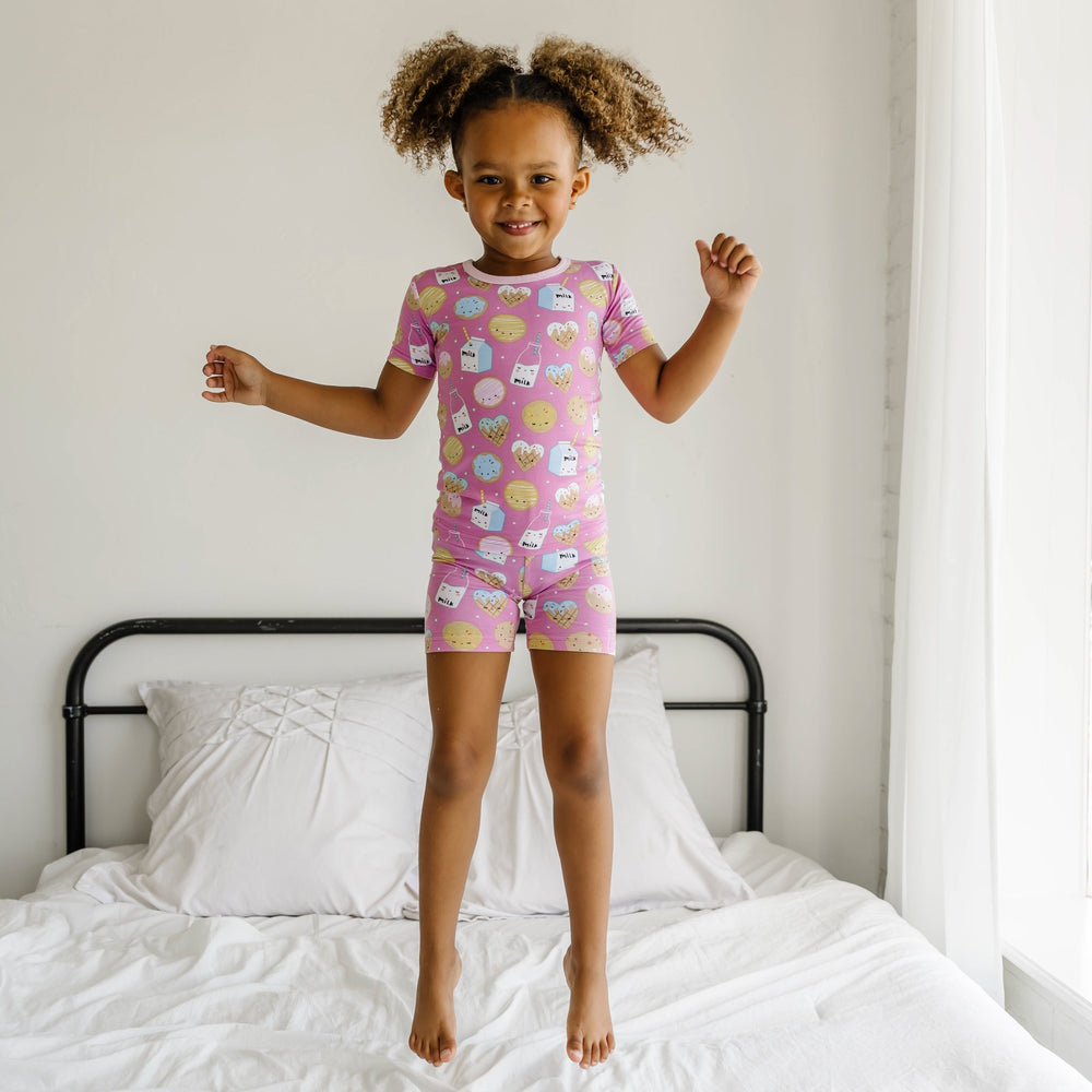 Click to see full screen - Image of little girl jumping on a bed. She is shown wearing a two-piece short sleeve and shorts pajama set in cookies and milk print. This print features milk cartons, colorful sprinkled cookies, and chocolate chip cookies that sit upon a pink background 