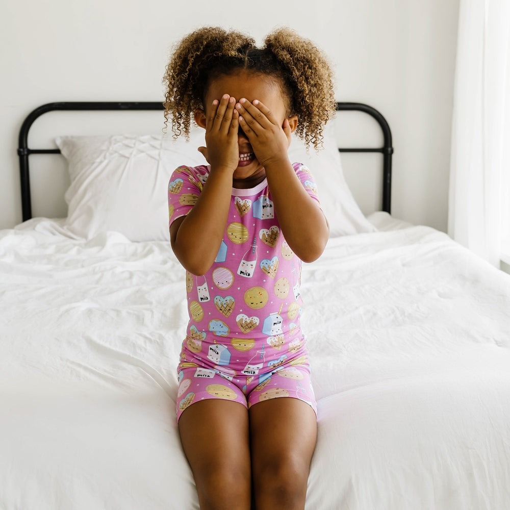 Image of little girl posing with her hands covering her eyes. She is shown wearing a two-piece short sleeve and shorts pajama set in cookies and milk print. This print features milk cartons, colorful sprinkled cookies, and chocolate chip cookies that sit 