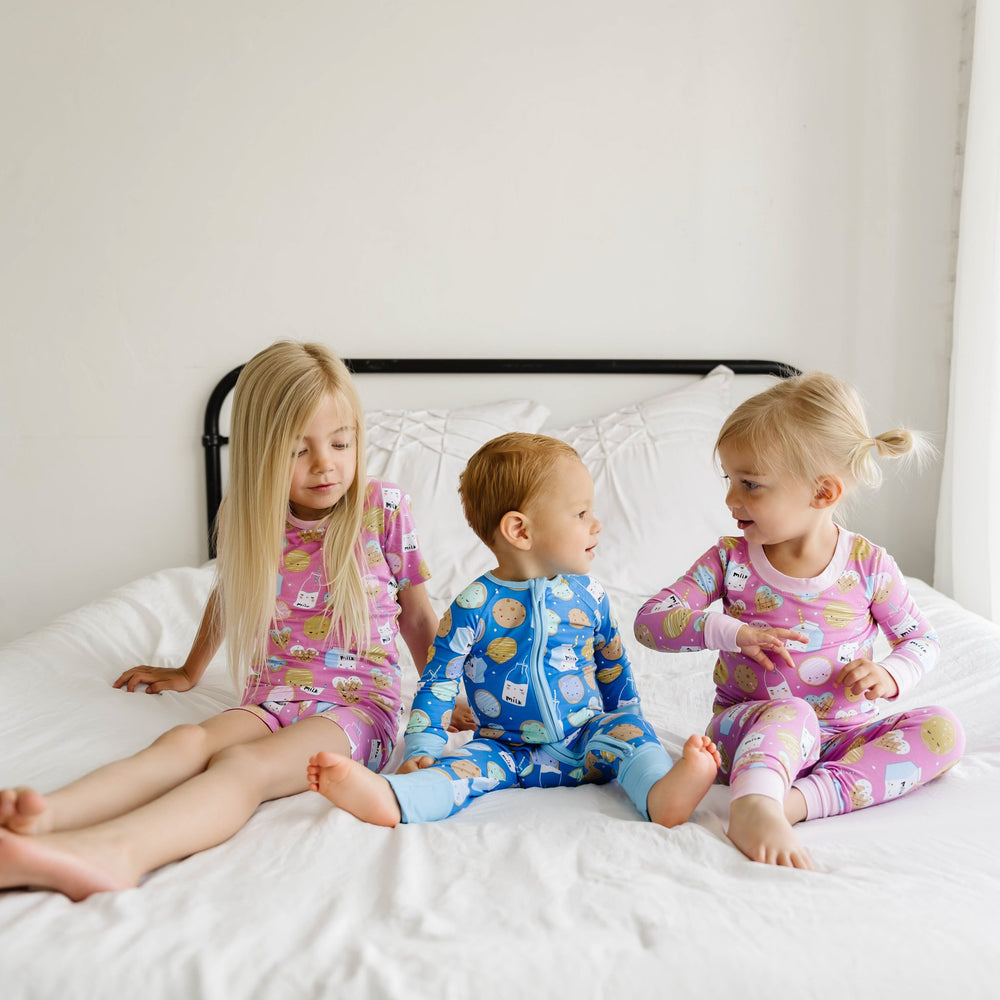 Image of three kids wearing matching pajamas in cookies and milk print. The two little girls are shown wearing the pajamas in pink, and the little boy is shown wearing blue.
