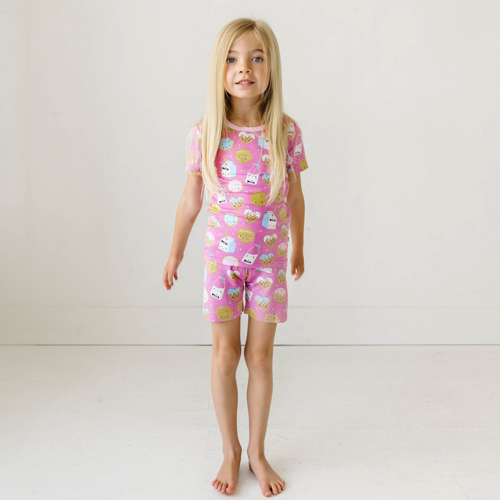Image of little girl wearing a two-piece short sleeve and shorts pajama set in cookies and milk print. This print features milk cartons, colorful sprinkled cookies, and chocolate chip cookies that sit upon a pink background with light pink trim. 