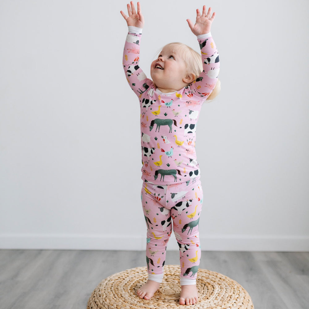 Image of little girl posing with her hands in the air. She is shown wearing a two-piece pajama set in a pink farm animals print. This print includes a pink background with white trim details. The farm animals featured on this print include cows, pigs, duc