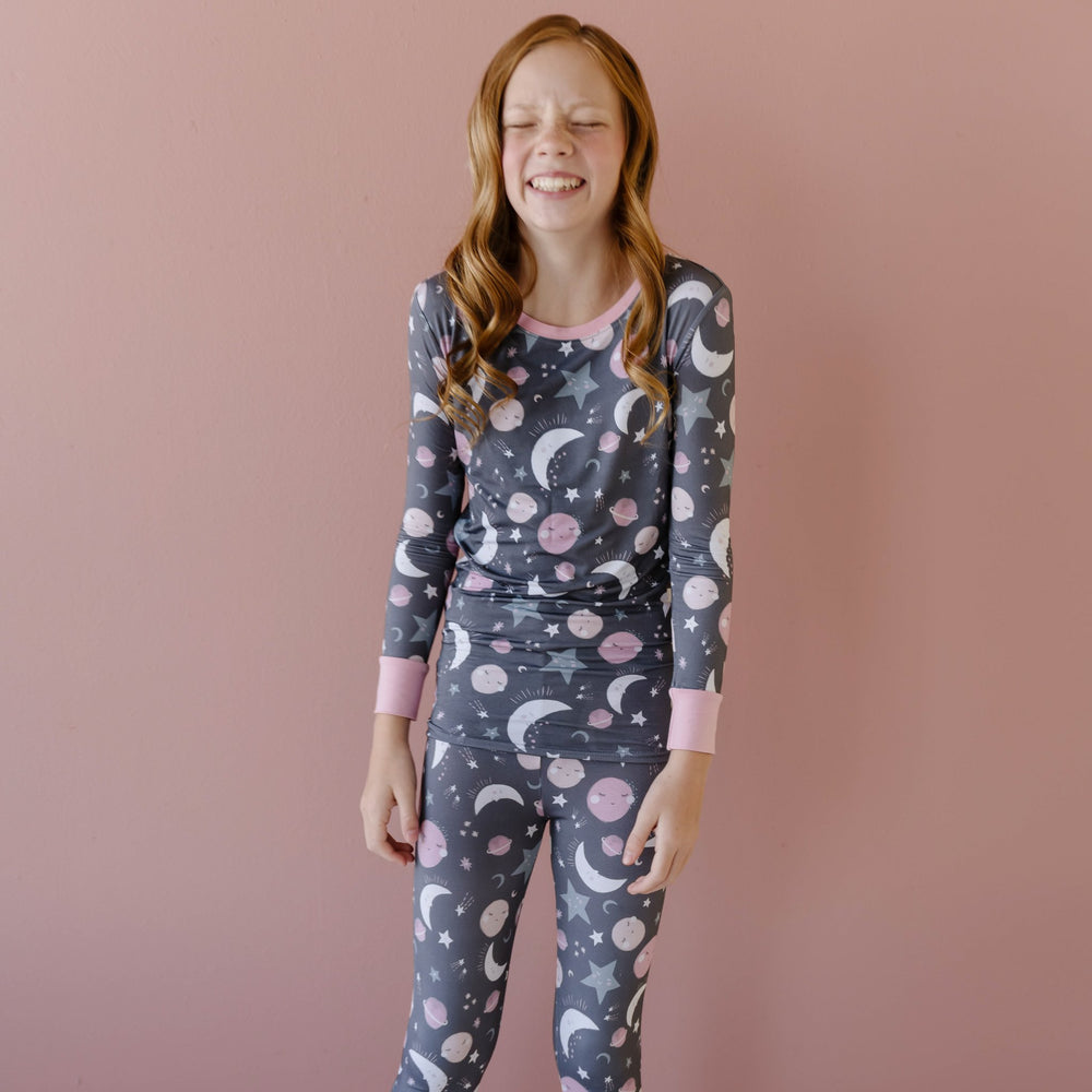 Image of little girl wearing a two-piece pajama set in pink to the moon and back print. This print features pink and gray moons, stars, and planets on a charcoal background with a matching pink trim. 