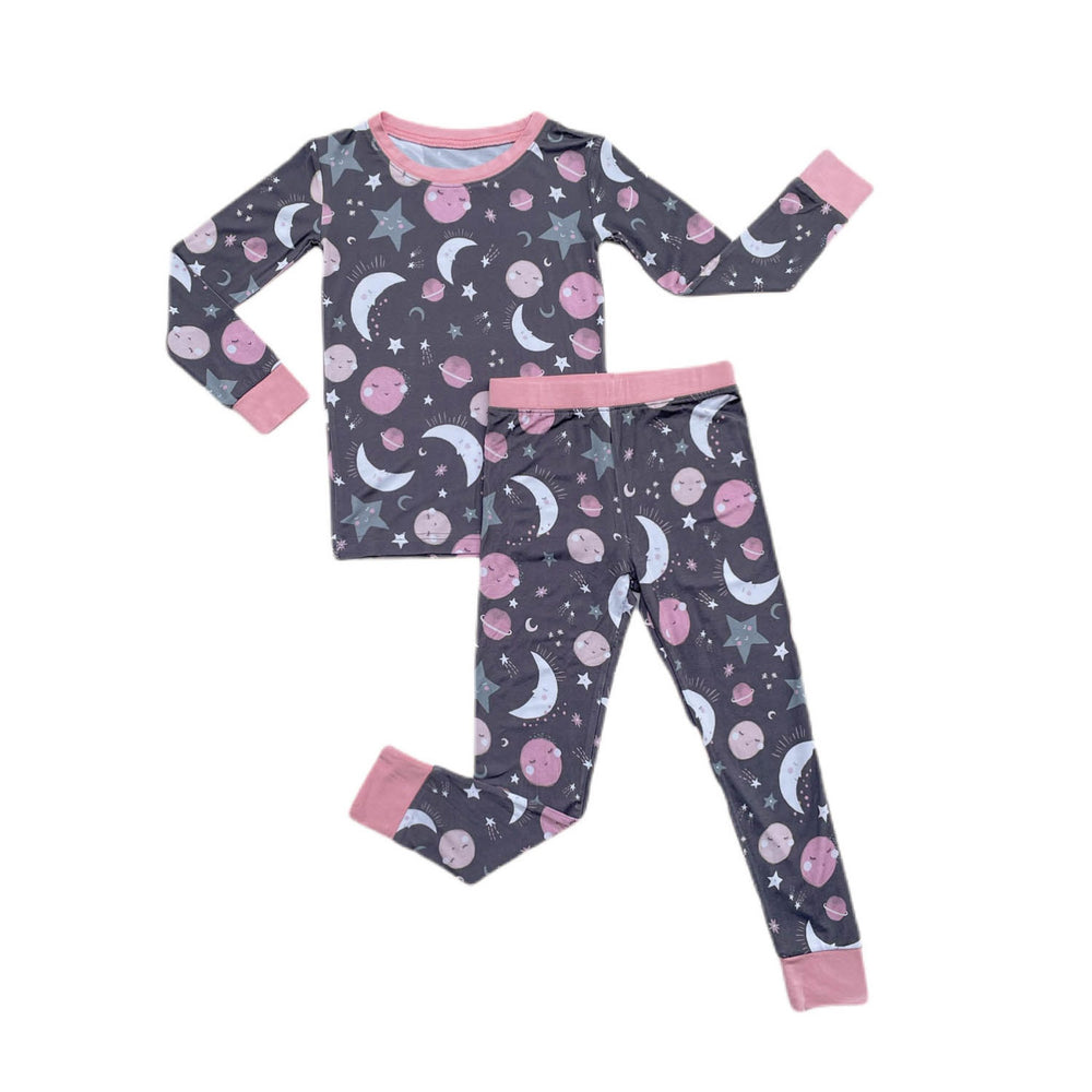 Flat lay image of two-piece pajama set in pink to the moon and back print. This print features pink and gray moons, stars, and planets on a charcoal background with a matching pink trim. 