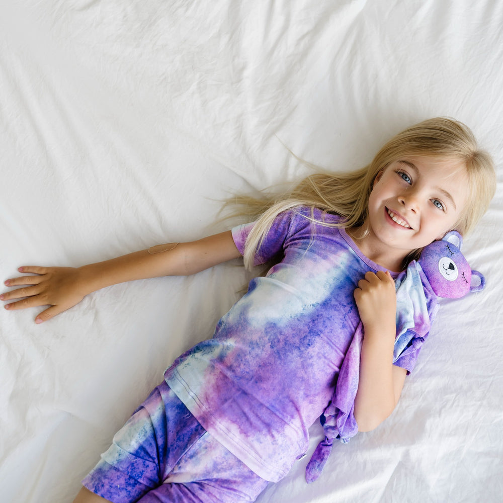 Image of girl laying down while wearing short sleeve and shorts pajama set in purple watercolor print and cuddling her bear lovey in Purple Watercolor print. This watercolor print includes shades of purple, hues of white, and the slightest hint of blue. 