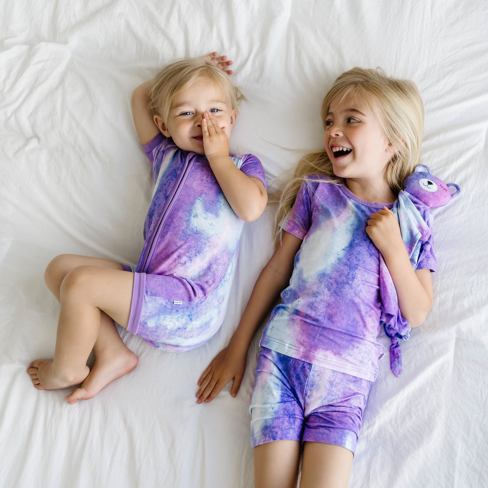 Image of sibling girls wearing coordinating purple watercolor printed shorty zippy and short sleeve and shorts pajama set. This watercolor print includes shades of purple, hues of white, and the slightest hint of blue. It is accented with purple trim.