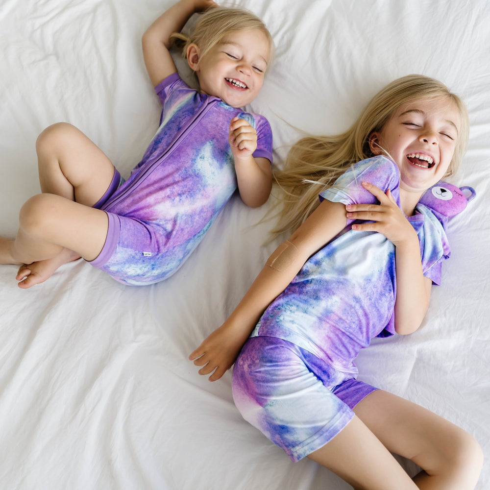 Image of sibling girls wearing coordinating purple watercolor printed shorty zippy and short sleeve and shorts pajama set. This watercolor print includes shades of purple, hues of white, and the slightest hint of blue. It is accented with purple trim.
