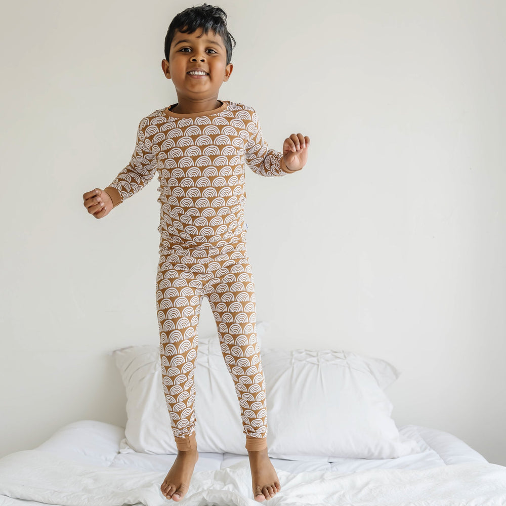 Click to see full screen - Image of little boy wearing a two-piece pajama set, featuring long sleeves and matching pants in Rust Rainbows print. This print features white rainbows that sit upon a rust brown background with matching rust brown trim.