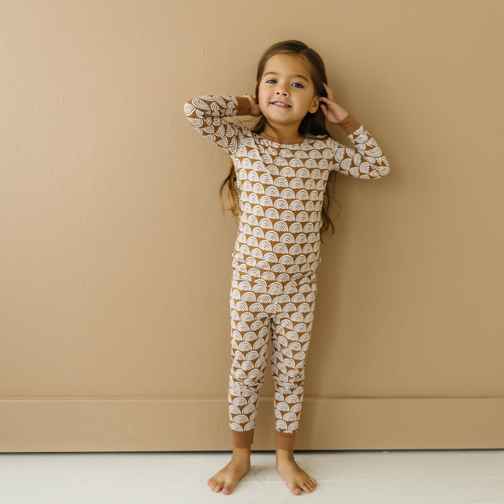 Image of little girl wearing a two-piece pajama set, featuring long sleeves and matching pants in Rust Rainbows print. This print features white rainbows that sit upon a rust brown background with matching rust brown trim