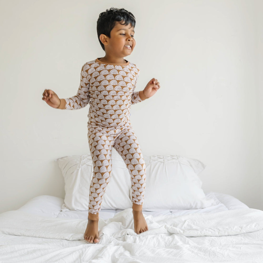 Click to see full screen - Image of little boy wearing a two-piece pajama set, featuring long sleeves and matching pants in Rust Rainbows print. This print features white rainbows that sit upon a rust brown background with matching rust brown trim