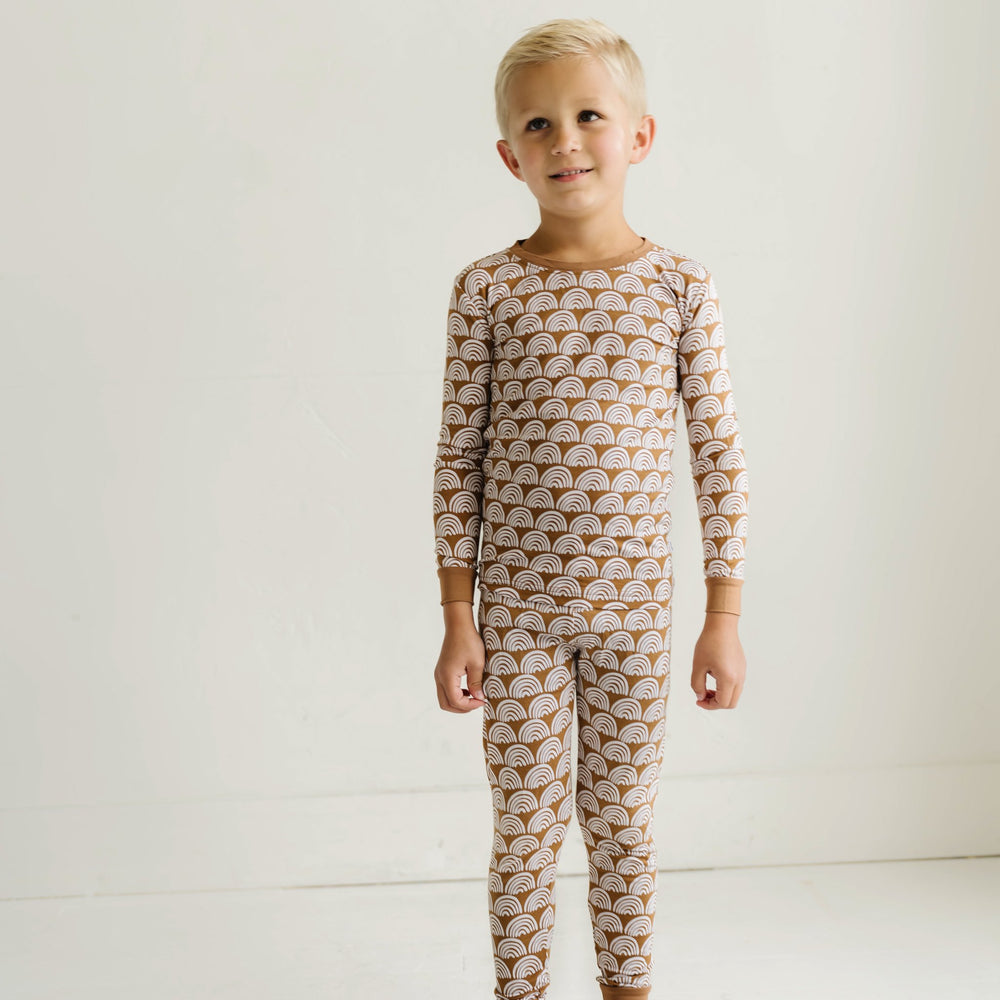 Image of little boy wearing a two-piece pajama set, featuring long sleeves and matching pants in Rust Rainbows print. This print features white rainbows that sit upon a rust brown background with matching rust brown trim