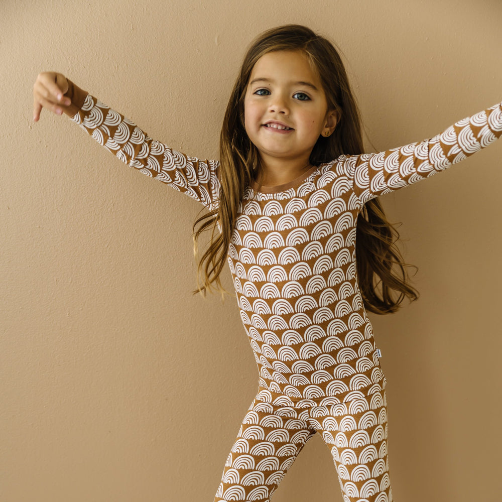 Click to see full screen - Image of little girl wearing a two-piece pajama set, featuring long sleeves and matching pants in Rust Rainbows print. This print features white rainbows that sit upon a rust brown background with matching rust brown trim