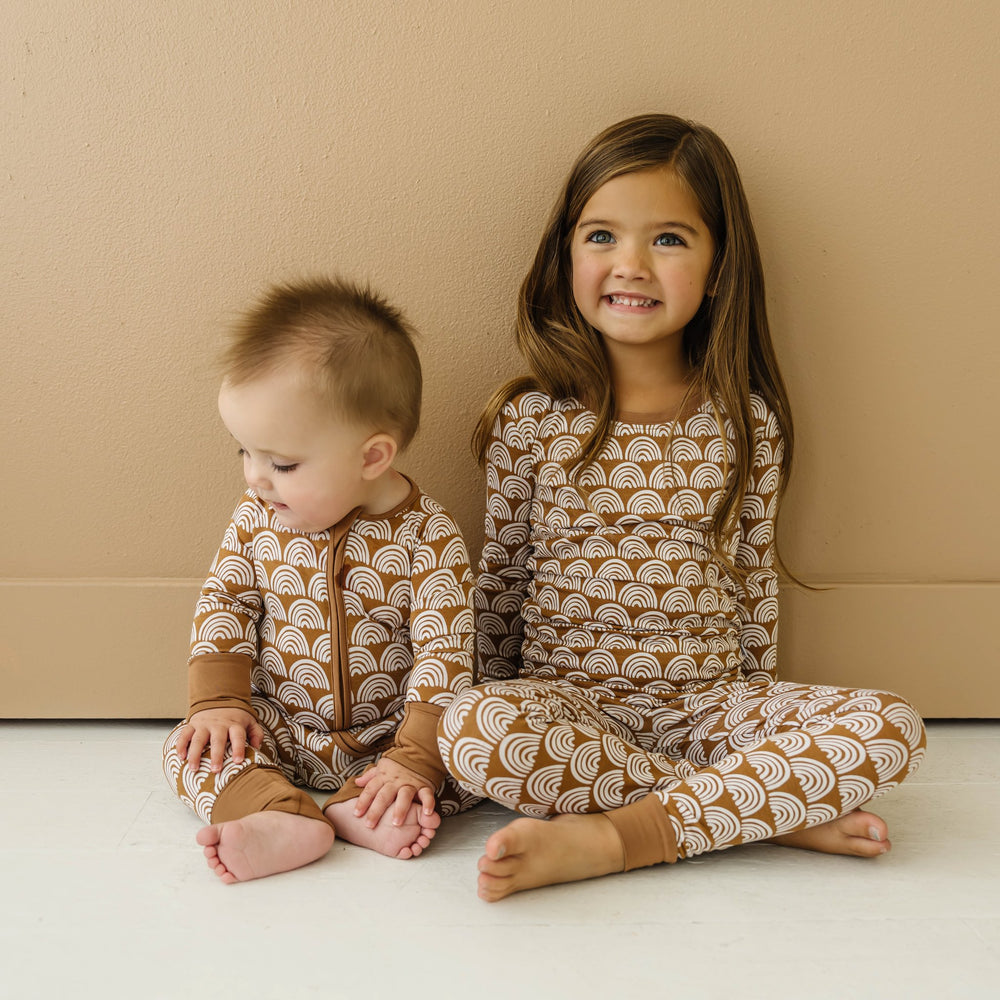 Image of infant baby and little girl wearing matching pajamas in Rust Rainbows print. The infant is shown wearing a long sleeve zip up romper, while the little girl is shown in a two-piece pajama set, featuring long sleeves and matching pants. This print features white rainbows that sit upon a rust brown background with matching rust brown trim.