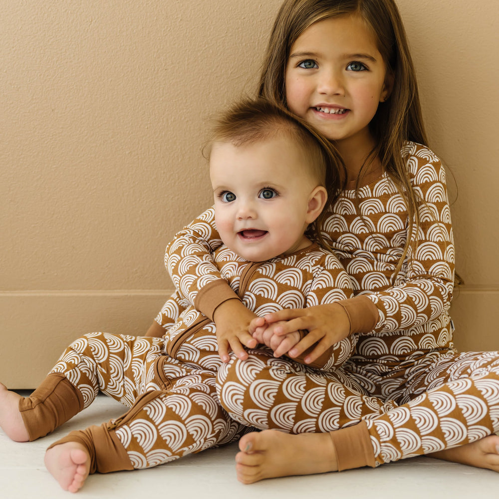 Image of infant baby and little girl wearing matching pajamas in Rust Rainbows print. The infant is shown wearing a long sleeve zip up romper, while the little girl is shown in a two-piece pajama set, featuring long sleeves and matching pants. This print features white rainbows that sit upon a rust brown background with matching rust brown trim.