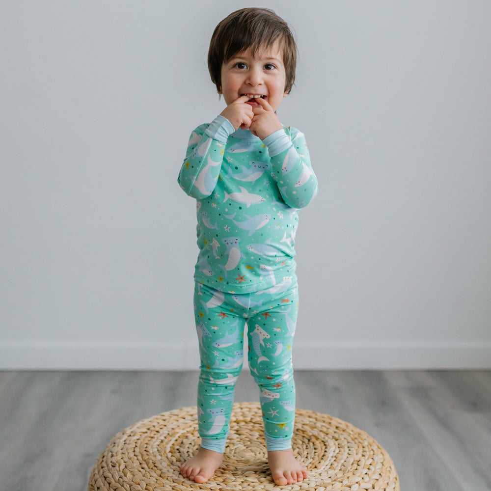 Click to see full screen - Image of toddler boy standing on top of a rattan pouf. He is shown wearing a two-piece pajama set with a shark print. This print includes hammerhead and great white sharks, featured on an aqua background with a light blue trim. 