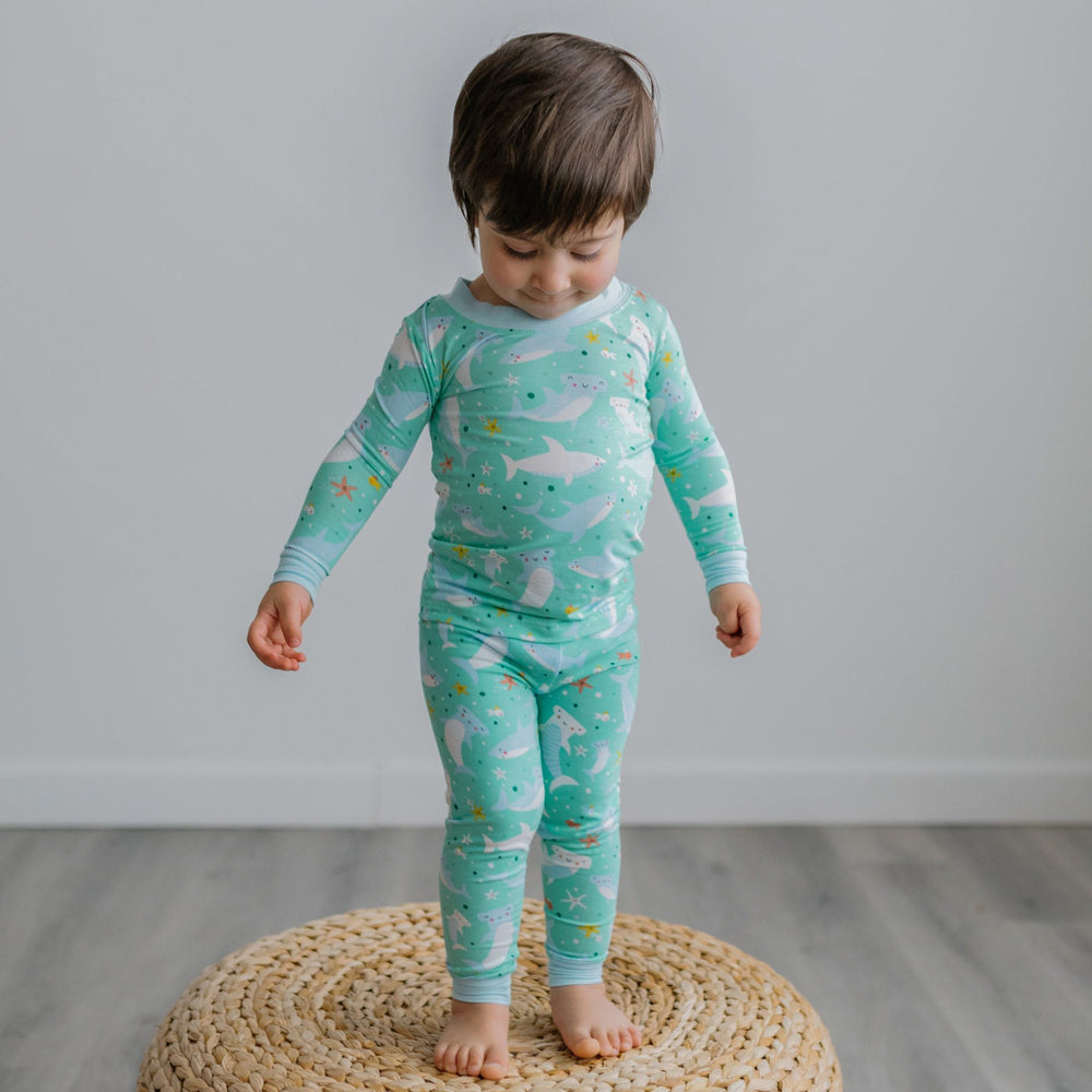 Image of toddler boy standing on top of a rattan pouf. He is shown wearing a two-piece pajama set with a shark print. This print includes hammerhead and great white sharks, featured on an aqua background with a light blue trim.