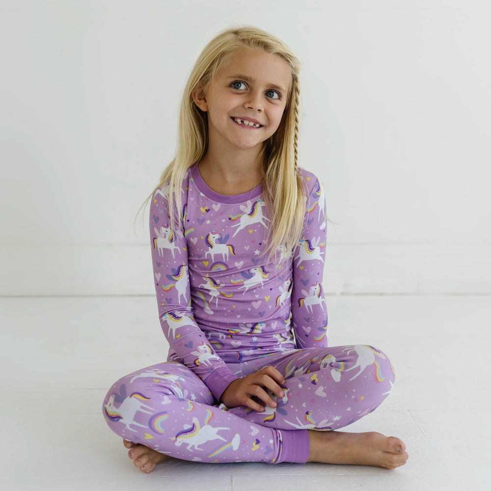 Click to see full screen - Image of little girl wearing a two-piece pajama set featuring long sleeves and matching long pants, in Sienna's Unicorns print. Flying unicorns with rainbow-colored manes gallop across a purple background with hearts, stars, and rainbows in this magical print.