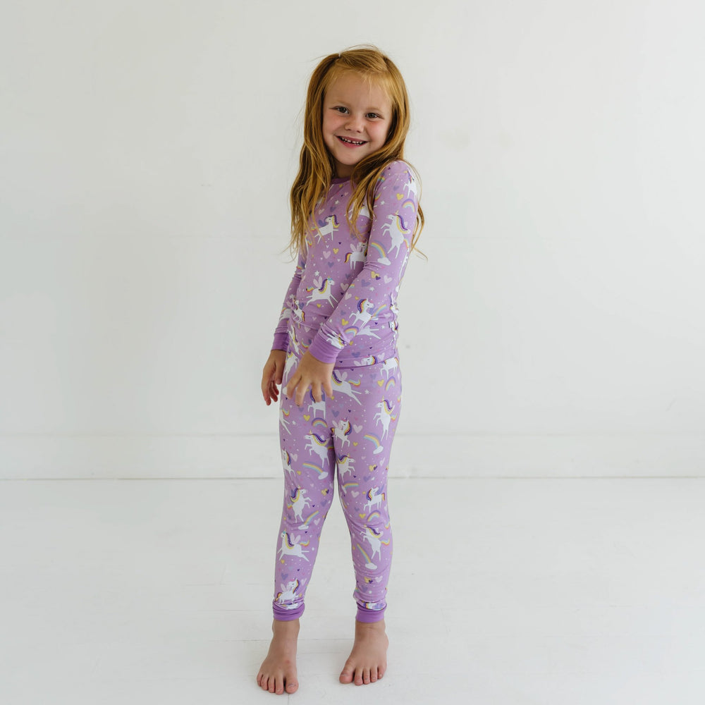 Image of little girl wearing a two-piece pajama set featuring long sleeves and matching long pants, in Sienna's Unicorns print. Flying unicorns with rainbow-colored manes gallop across a purple background with hearts, stars, and rainbows in this magical print.