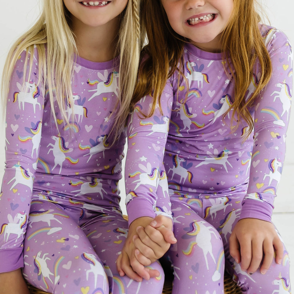 Image of two little girl swearing matching two-piece pajama sets featuring long sleeves and matching long pants, in Sienna's Unicorns print. Flying unicorns with rainbow-colored manes gallop across a purple background with hearts, stars, and rainbows in this magical print.
