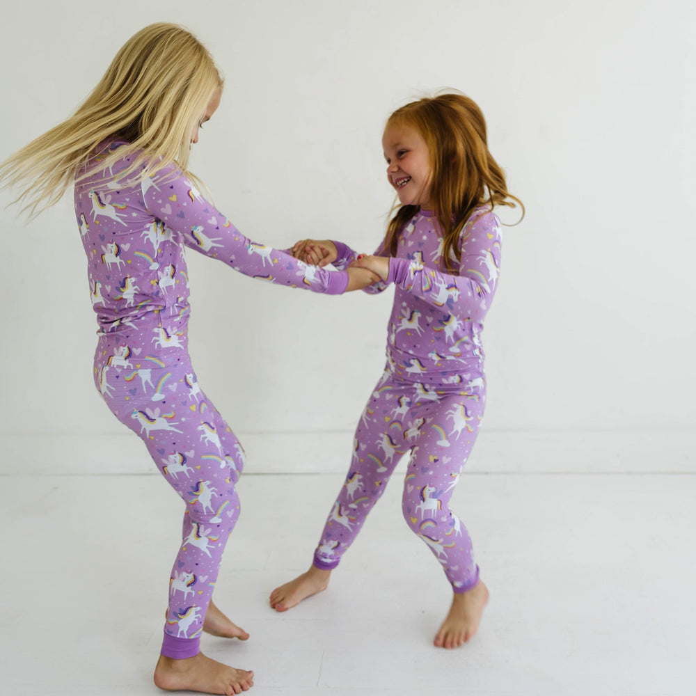 Image of two little girl swearing matching two-piece pajama sets featuring long sleeves and matching long pants, in Sienna's Unicorns print. Flying unicorns with rainbow-colored manes gallop across a purple background with hearts, stars, and rainbows in this magical print.