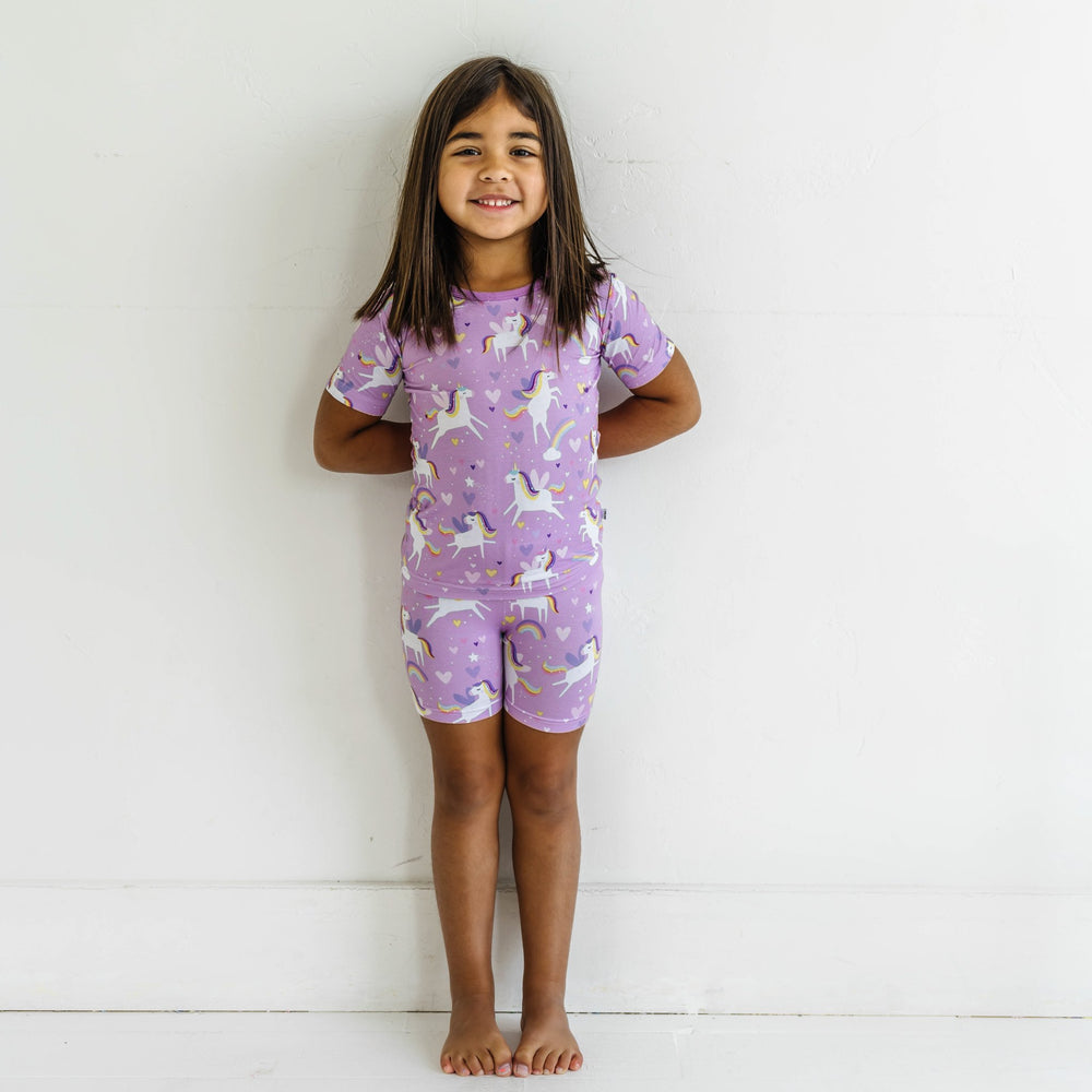 Click to see full screen - Image of little girl wearing a two-piece short sleeve and shorts pajama set in Sienna's Unicorns print. Flying unicorns with rainbow-colored manes gallop across a purple background with hearts, stars, and rainbows in this magical print.