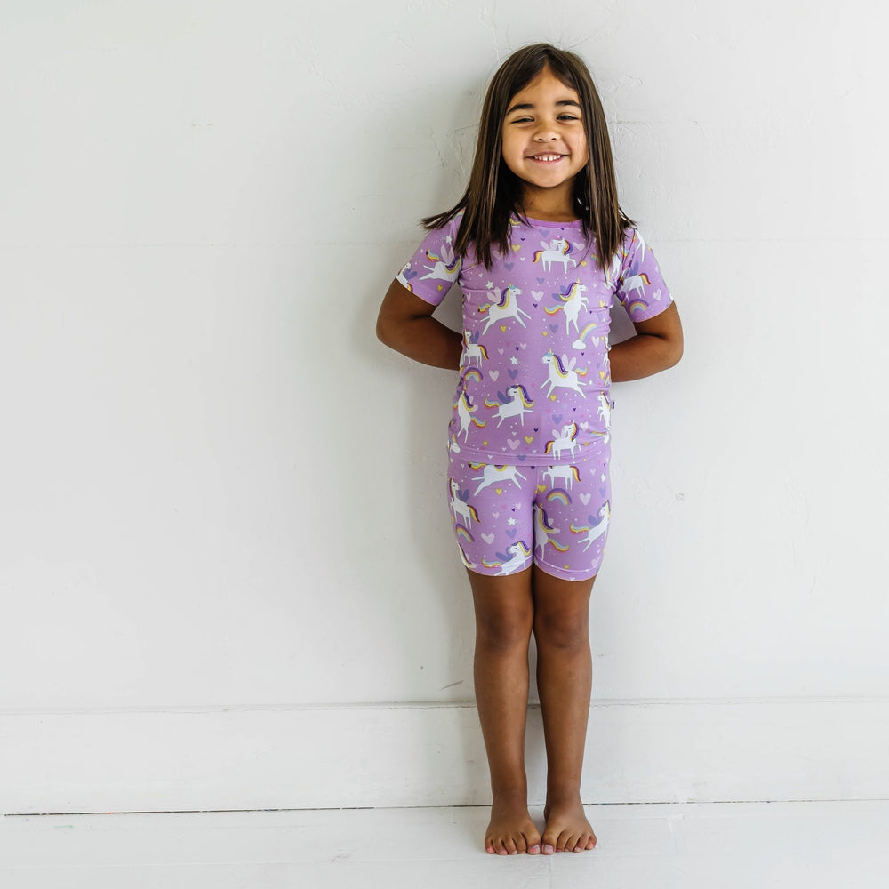 Click to see full screen - Image of little girl wearing a two-piece short sleeve and shorts pajama set in Sienna's Unicorns print. Flying unicorns with rainbow-colored manes gallop across a purple background with hearts, stars, and rainbows in this magical print.