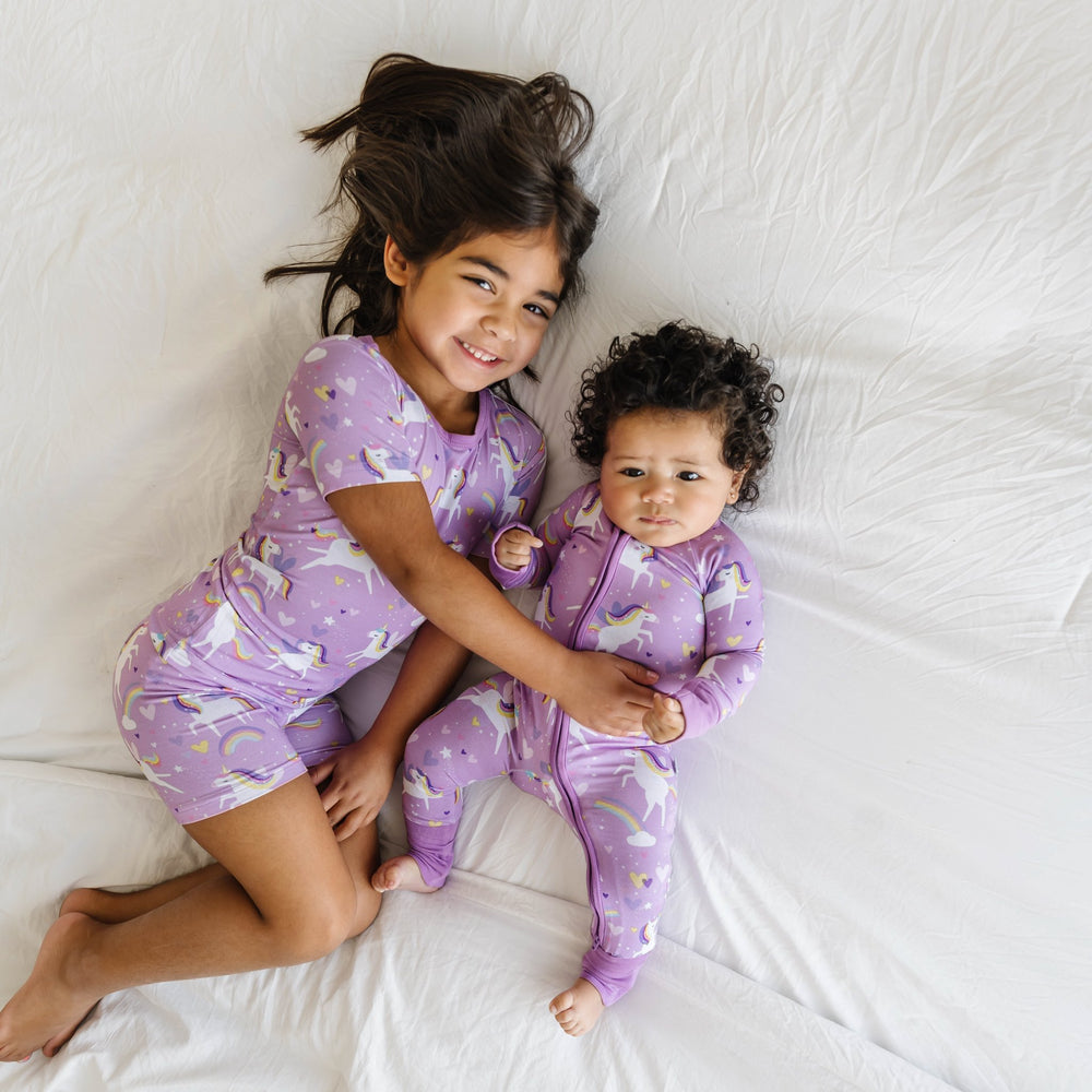 Click to see full screen - Image of two little girls wearing matching unicorn printed pajama sets. The bigger sister is wearing a short sleeve and shorts pajama set and the little sister is shown wearing a zip up romper. This print features flying unicorns with rainbow-colored manes gallop across a purple background with hearts, stars, and rainbows