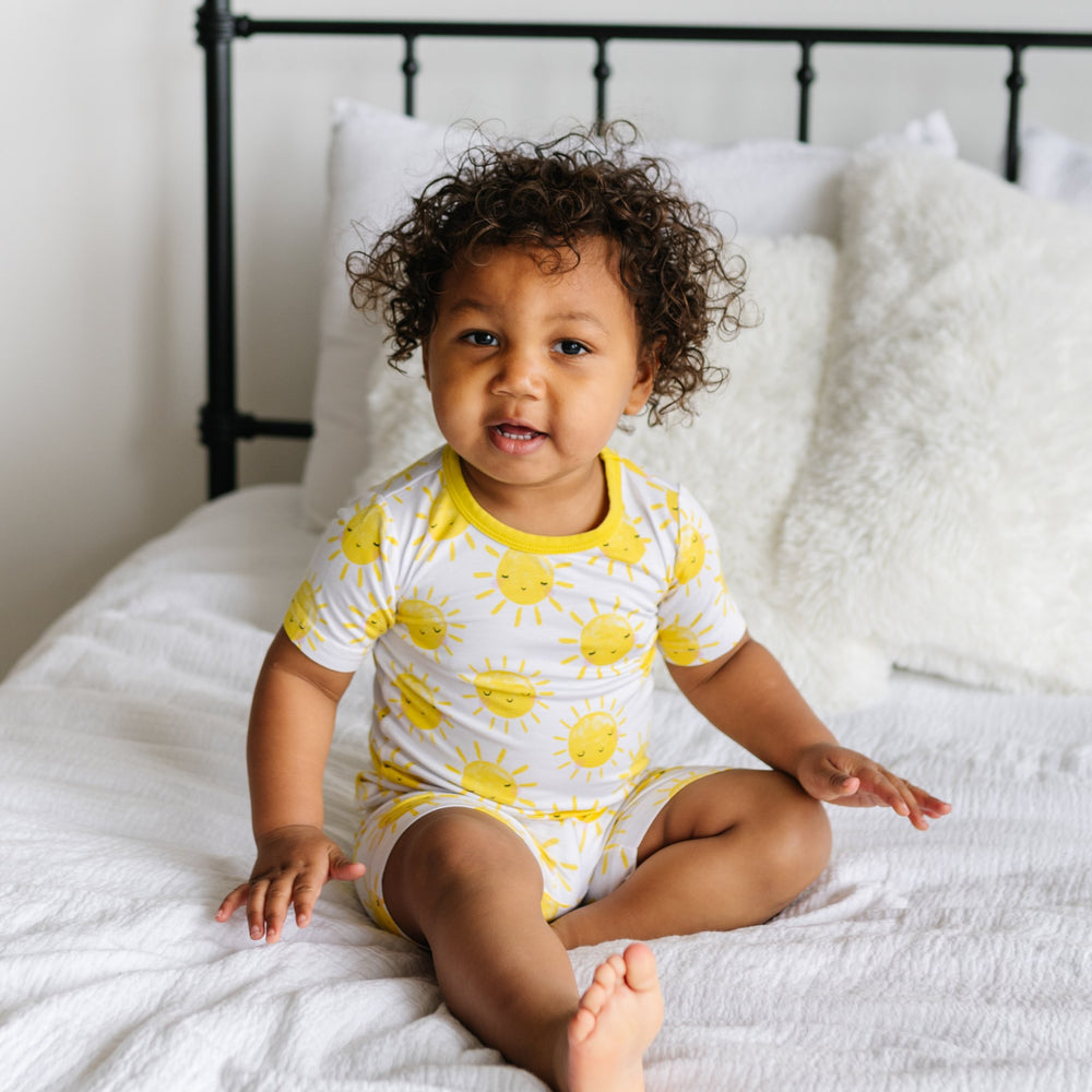 Click to see full screen - Image of toddler sitting on a bed wearing a two-piece short sleeve and shorts pajama set in Sunshine print. The yellow smiling suns on this print sit upon a white background, accented with yellow trim.