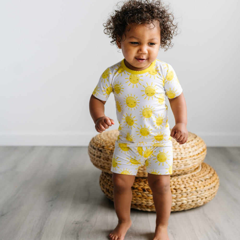 Image of toddler wearing a two-piece short sleeve and shorts pajama set in Sunshine print. The yellow smiling suns on this print sit upon a white background, accented with yellow trim.