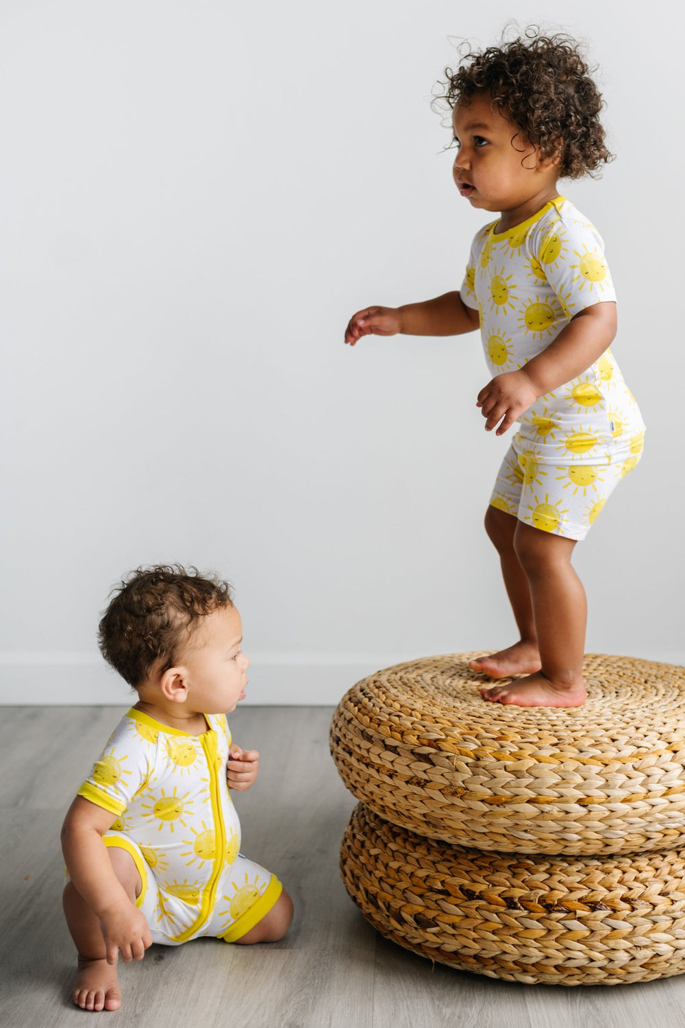 Click to see full screen - Image of two kids wearing matching sunshine printed pajamas. The yellow smiling suns on this print sit upon a white background, accented with yellow trim.