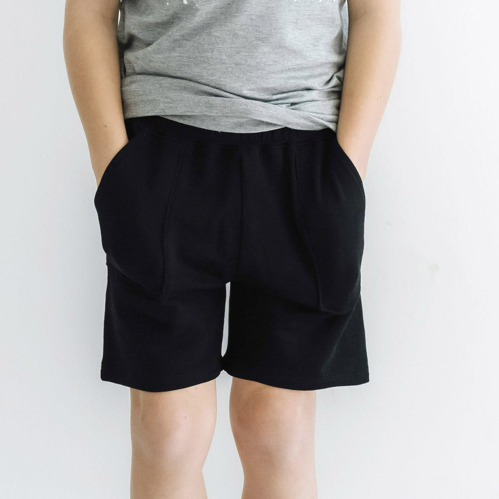 Close up image of a child posing with their hands in their pockets wearing Black shorts 
