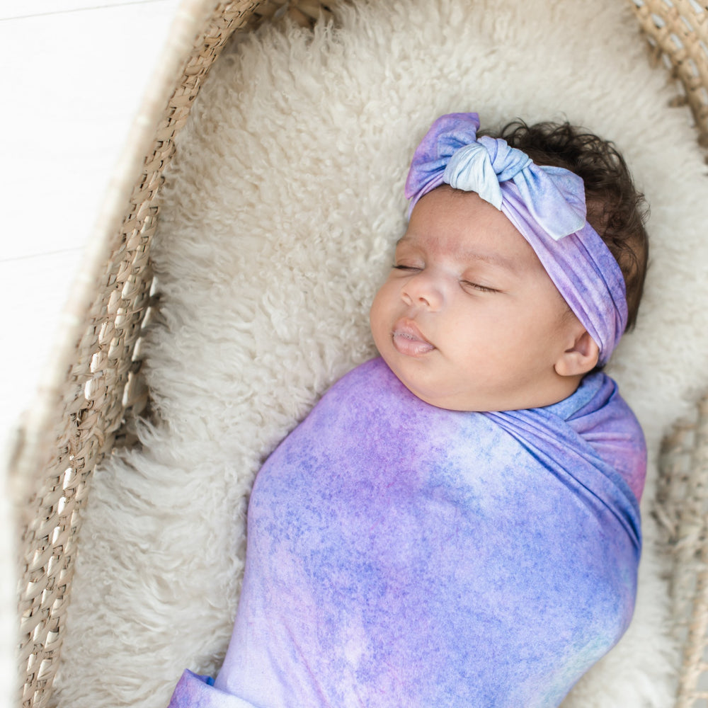 Image of infant girl laying in bassinet basket wearing swaddle and headband set in purple watercolor print. This watercolor print includes shades of purple, hues of white, and the slightest hint of blue.