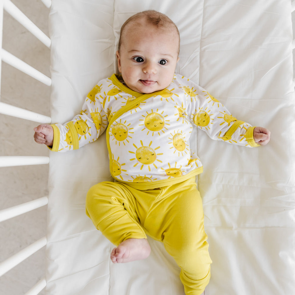 Image of an infant wearing a two-piece crossover set that features a long sleeve wrap style top with snap closures and coordinating pants with convertible footies. This style is in the Sunshine print that features sweet yellow sunshines on a white brown background with matching yellow trim.