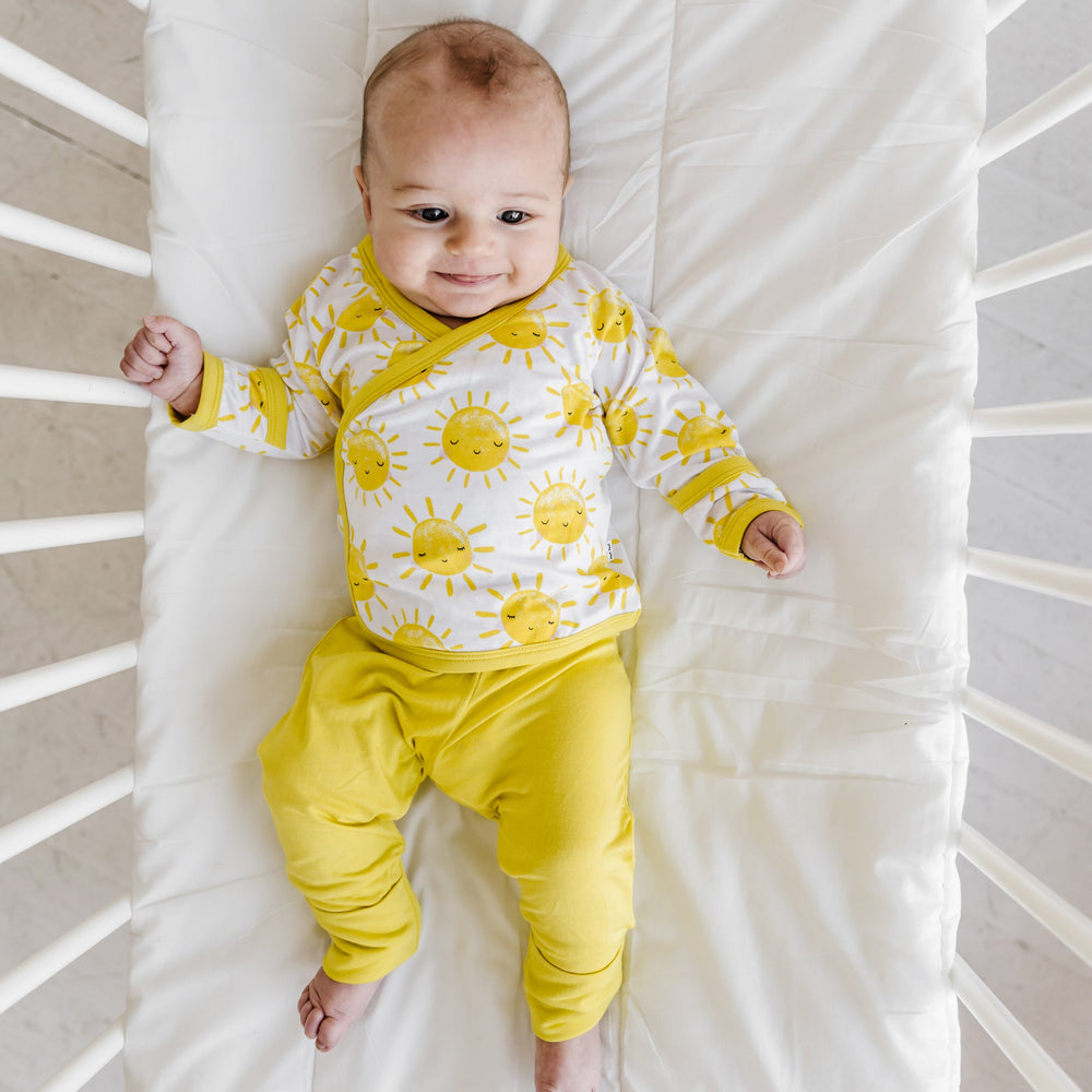 Image of an infant wearing a two-piece crossover set that features a long sleeve wrap style top with snap closures and coordinating pants with convertible footies. This style is in the Sunshine print that features sweet yellow sunshines on a white brown background with matching yellow trim.