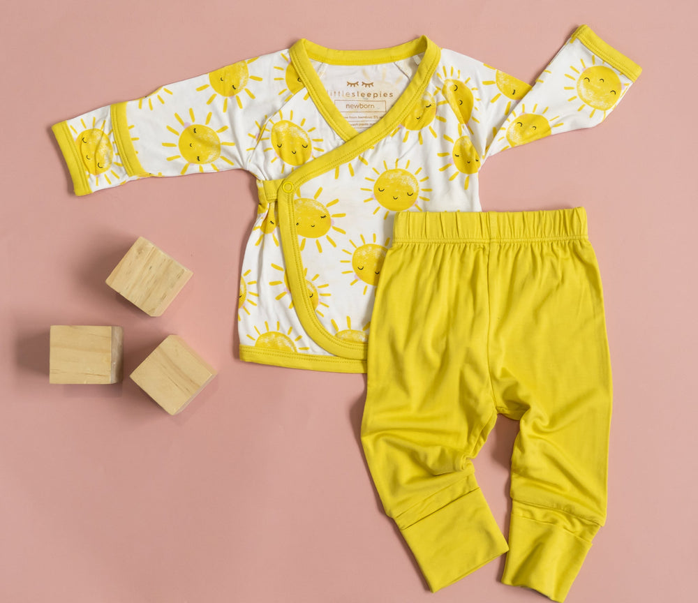 Flat lay image of a two-piece crossover set that features a long sleeve wrap style top with snap closures and coordinating pants with convertible footies. This style pictured has the wrap style top in Sunshine print with sweet yellow sunshines on a white brown background with matching yellow trim.